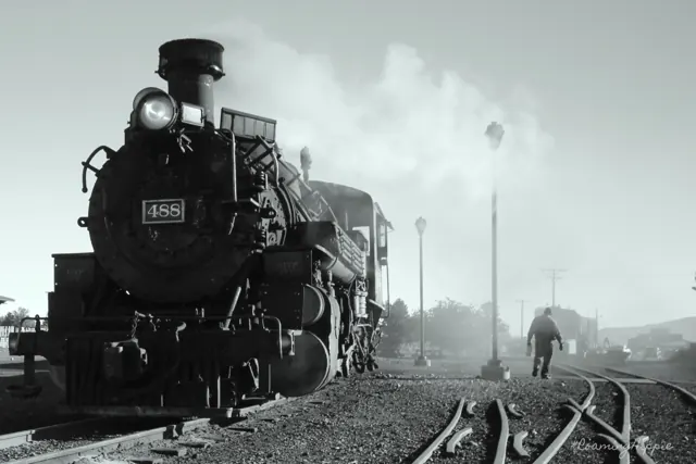 A black and white steam locomotive letting off steam in a rail yard